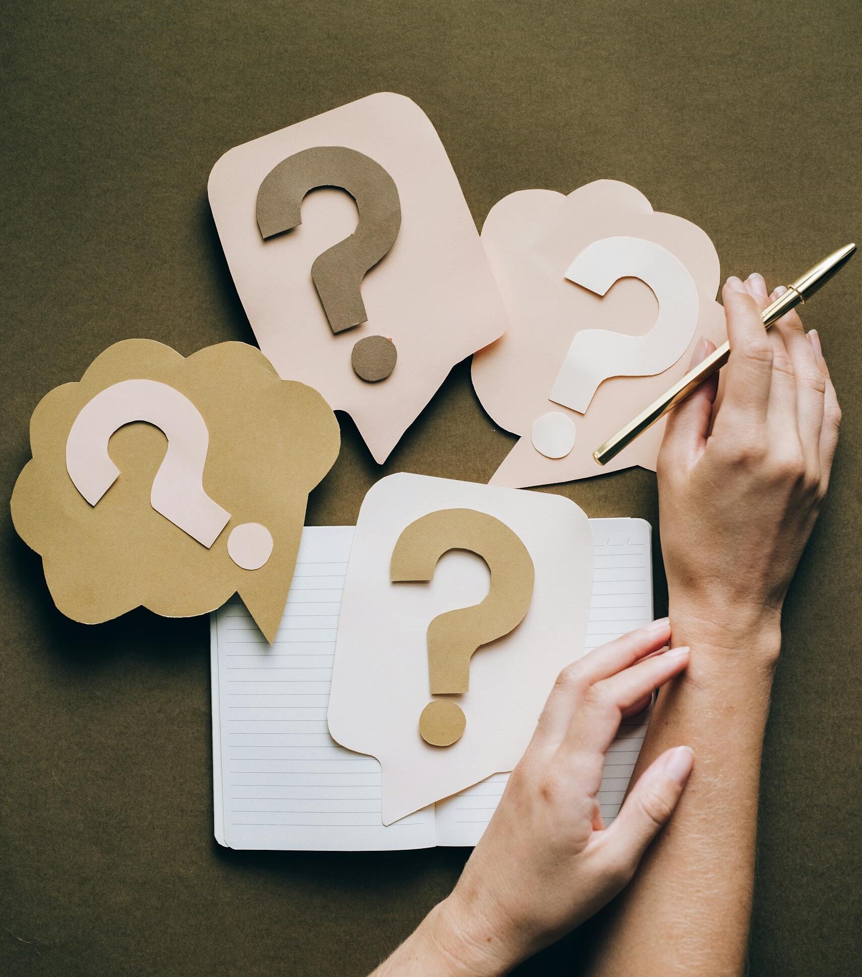Hands holding pen next to paper question marks on notebook, symbolizing inquiry and creativity.